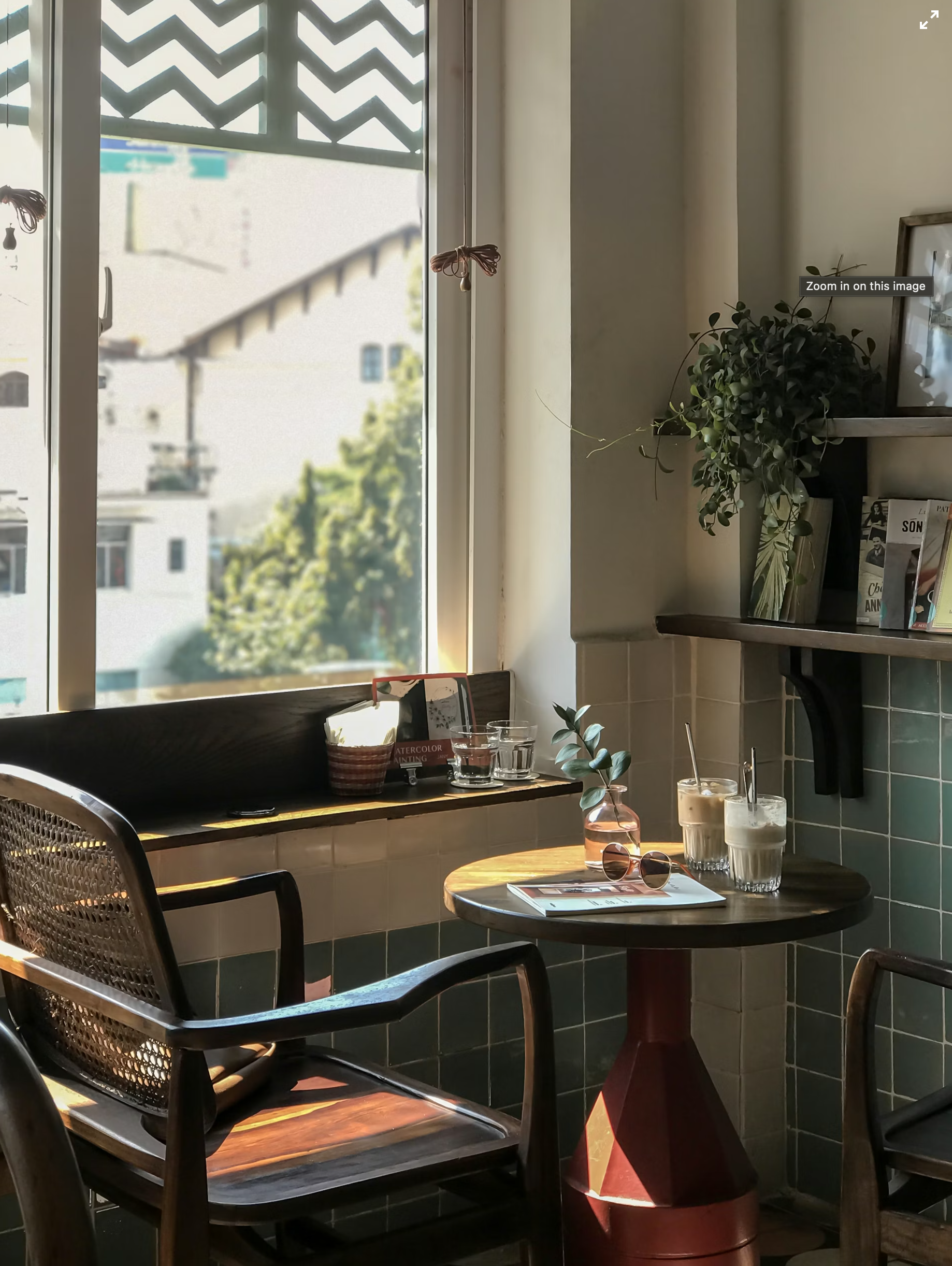 Sunlit café window table with two coffees, a magazine, and a rattan chair