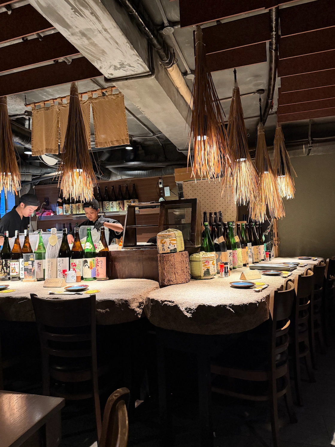 Mangetsu izakaya interior — stone counter, rows of sake bottles, straw lanterns hanging above