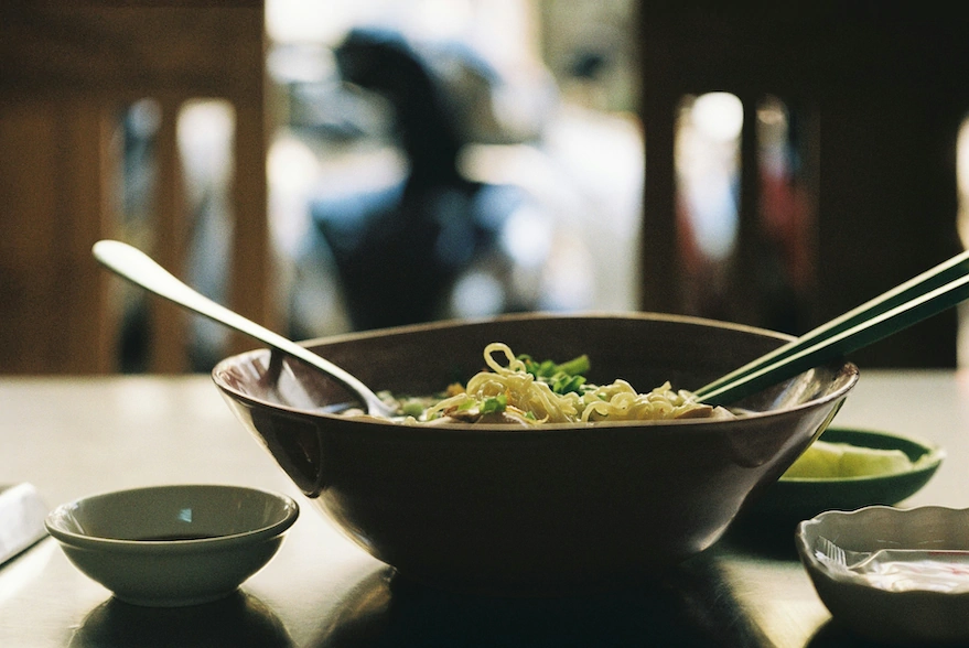 Bowl of phở with chopsticks and spoon, motorbike blurred through a shophouse window