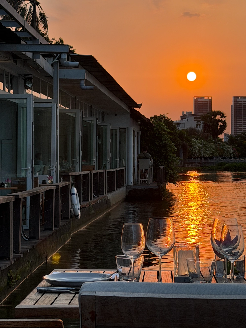 The Deck Saigon at sunset — riverside table with wine glasses, candle, and orange sun over the Saigon River